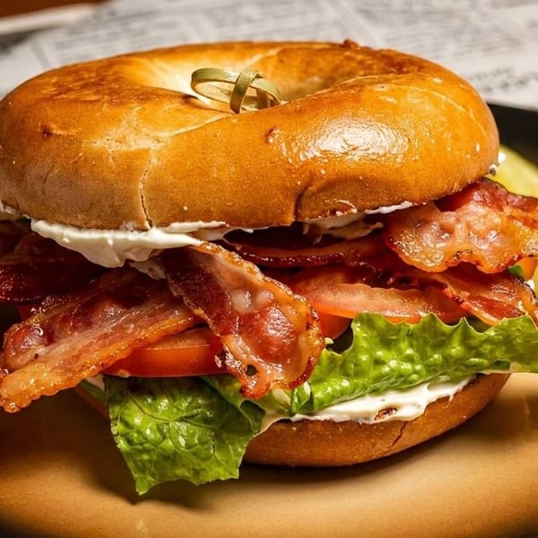 A bagel sandwich with bacon, lettuce, tomato, and cream cheese, served with a pickle spear.