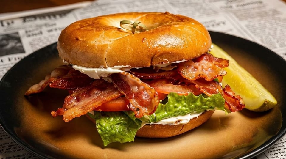 A bagel sandwich with bacon, lettuce, tomato, and cream cheese, served with a pickle spear.