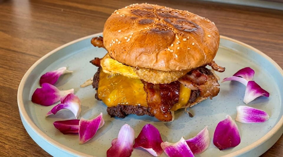 Gourmet burger with melted cheese, bacon, and a fried egg, garnished with purple flower petals.