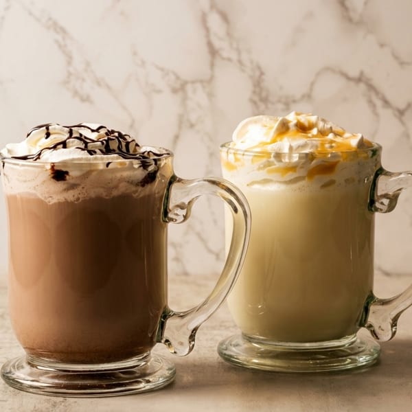 Two mugs of hot chocolate, one chocolate and one vanilla, topped with whipped cream and drizzle.
