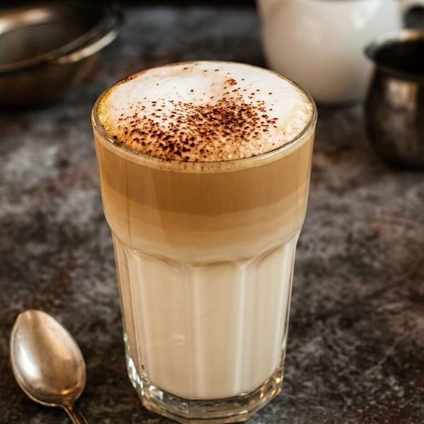 Tall glass of layered latte with frothy milk and cocoa powder, next to a spoon and creamers.