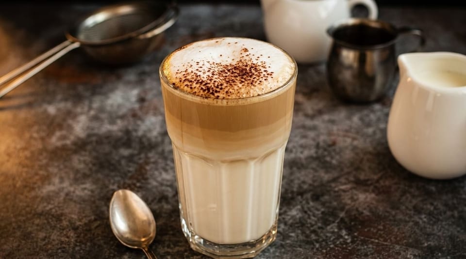 Tall glass of layered latte with frothy milk and cocoa powder, next to a spoon and creamers.