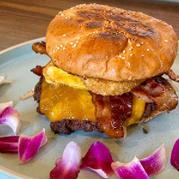 A gourmet burger with cheese, bacon, and a fried egg on a light blue plate with pink petals.